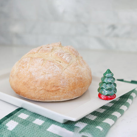 Loaf of bread on a white plate with a decorative Christmas tree figurine on a checkered cloth.