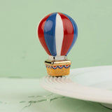 A small decorative hot air balloon charm or bead with red, white, and blue vertical stripes and a gold-toned basket, displayed on a white plate against a pale green background.