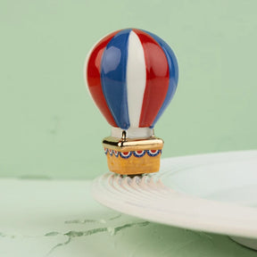 A small decorative hot air balloon charm or bead with red, white, and blue vertical stripes and a gold-toned basket, displayed on a white plate against a pale green background.