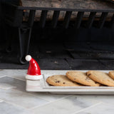 A white rectangular serving tray holding several chocolate chip cookies, with a small red and white Santa hat placed on the left side, set on a light stone surface near a fireplace.