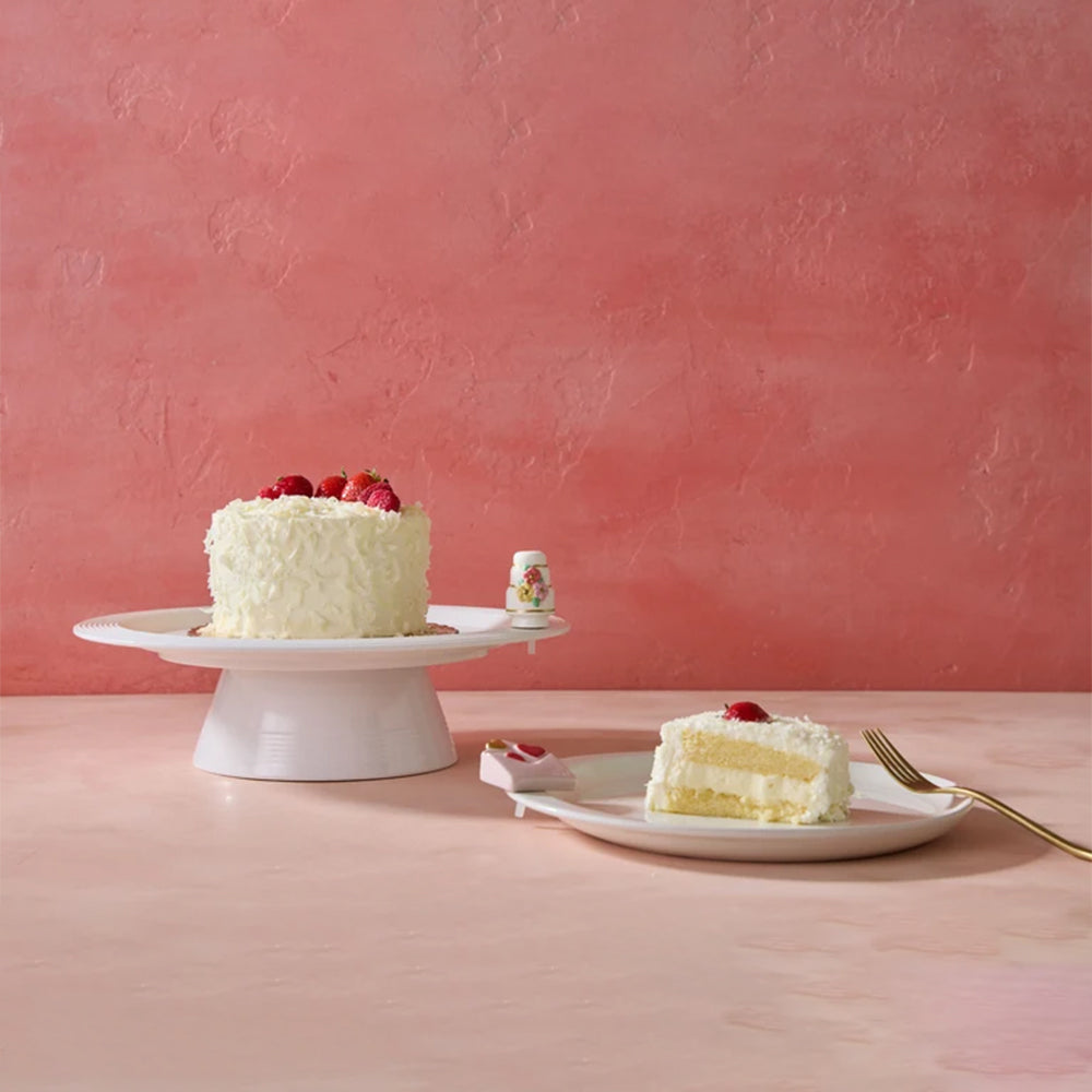 A white frosted cake with red berries sits on a white pedestal plate, while a slice of layered cream cake with a red berry on top rests on a white plate with a gold fork, set against a textured pink wall and matching pink surface.