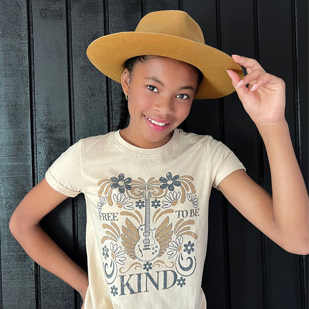 Child wearing a beige t-shirt with a graphic design and text, holding a brown hat against a dark wooden background.