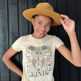 Child wearing a beige t-shirt with a graphic design and text, holding a brown hat against a dark wooden background.