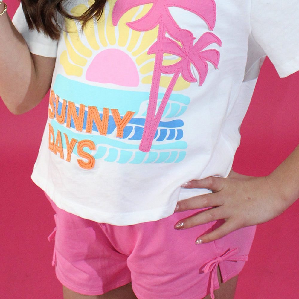 Person wearing a white t-shirt with 'Sunny Days' and palm tree design on a pink background