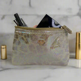 Iridescent makeup pouch with gold zipper and logo, containing cosmetic items on a marble surface.