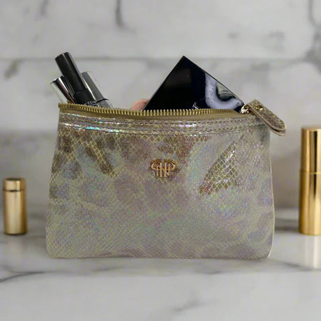 Iridescent makeup pouch with gold zipper and logo, containing cosmetic items on a marble surface.