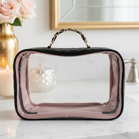 Clear makeup bag with black quilted handle on a vanity table with decorative items.
