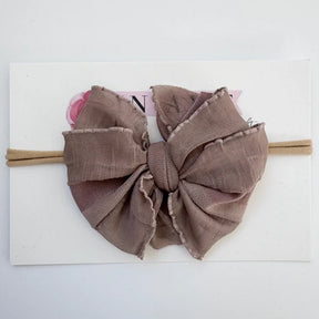A large fabric bow headband in a dusty taupe-brown color, displayed on a white backing card.