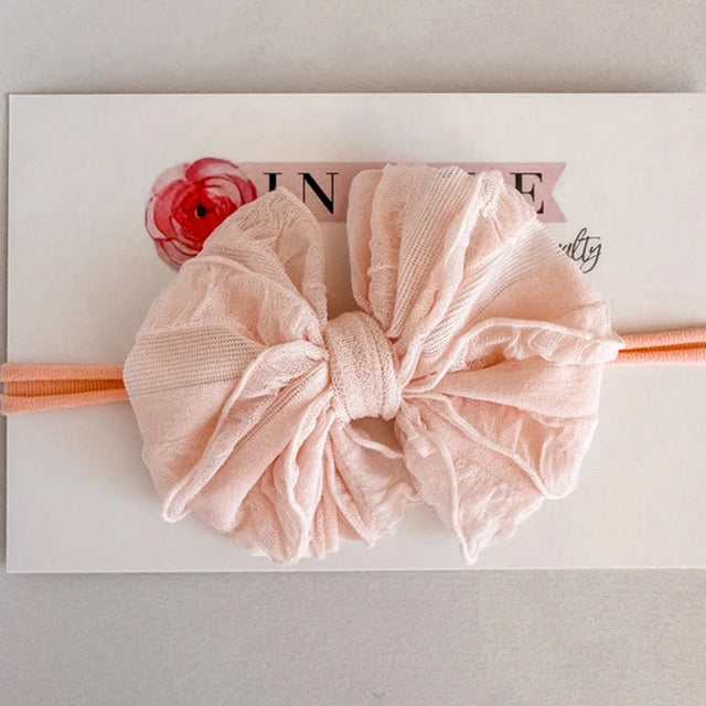 A large pale pink fabric bow headband with a matching pink elastic band, displayed on a white card with a red flower graphic.