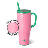 A pink insulated travel tumbler with a green handle, green lid trim, and green straw, shown on a white background. A circular inset highlights the golf-ball textured pattern, with the text 'FEELS LIKE A GOLF BALL' and the brand name 'swig' visible on the tumbler.