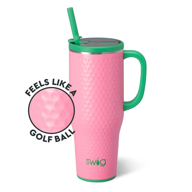 A pink insulated travel tumbler with a green handle, green lid trim, and green straw, shown on a white background. A circular inset highlights the golf-ball textured pattern, with the text 'FEELS LIKE A GOLF BALL' and the brand name 'swig' visible on the tumbler.