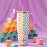 Colorful checkered tumbler with pink handle and straw against a purple background with geometric blocks.