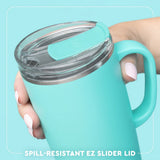 A close-up of a hand holding a mint green insulated tumbler with a clear lid and turquoise spill-resistant ez slider lid. The background is a solid light turquoise with a white rounded border and text at the bottom reading 'SPILL-RESISTANT EZ SLIDER LID'.