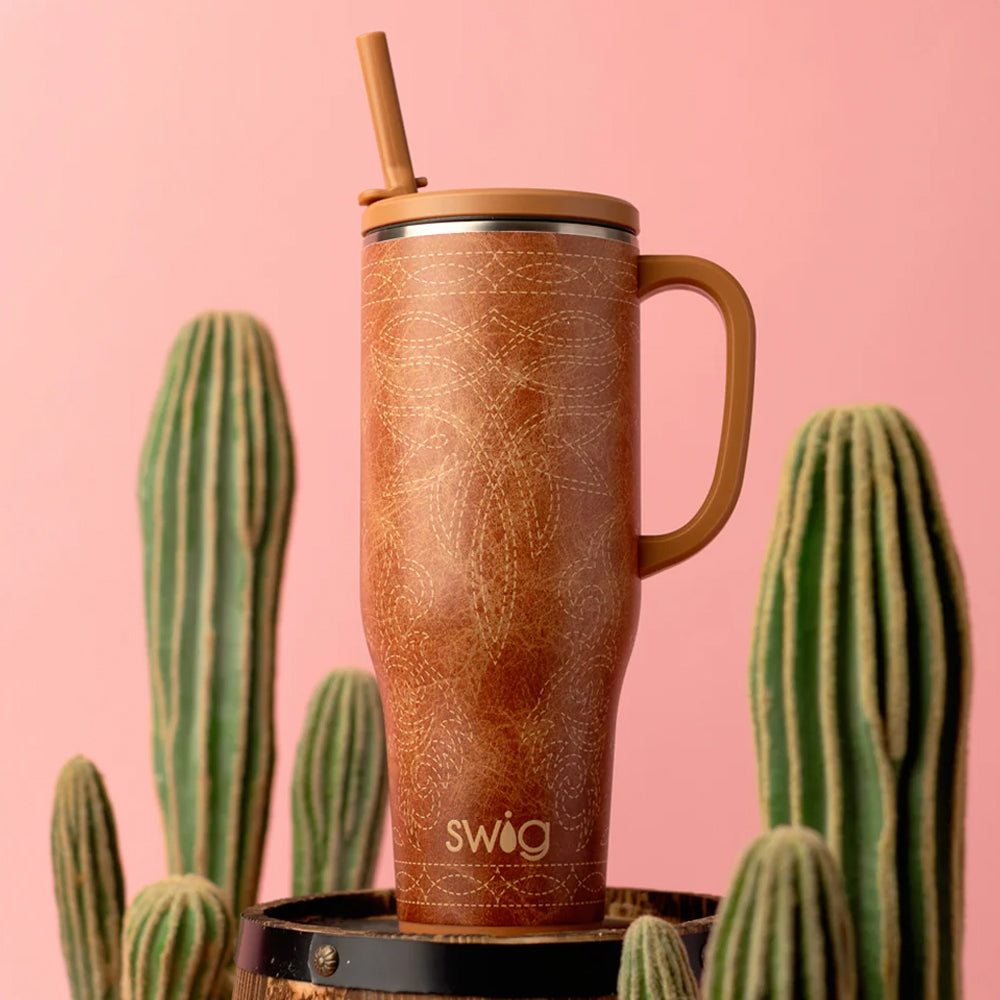 A tall handled tumbler with a bamboo straw and bamboo lid, featuring a brown textured pattern, displayed on a black barrel stand against a pink background with cacti.