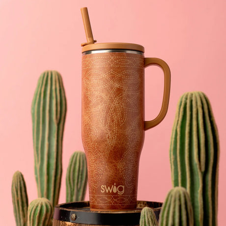 A tall handled tumbler with a bamboo straw and bamboo lid, featuring a brown textured pattern, displayed on a black barrel stand against a pink background with cacti.