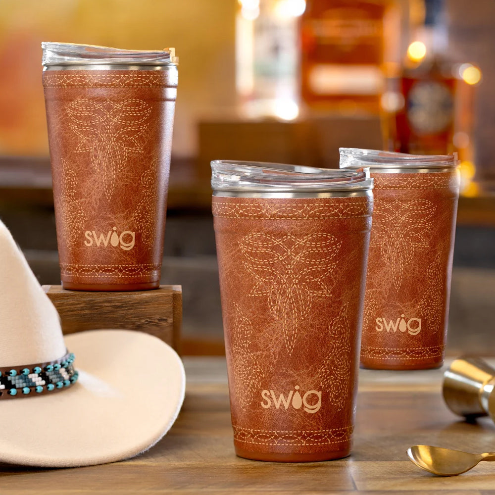 Three SWOOG insulated tumblers with clear lids are arranged on a wooden surface in a warm bar-like setting. A white cowboy hat with a turquoise band is partially visible on the left, and a gold spoon and metal container appear at the lower right.