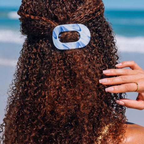 Person with curly hair wearing a blue and white hair tie against a blurred beach background