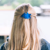 Person with blue hair clip in blonde hair, blurred outdoor background