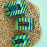 Three green and blue striped hair clips  on a woven mat with a light blue background