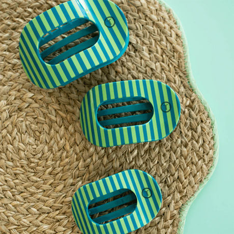 Three green and blue striped clips on a woven mat with a light blue background