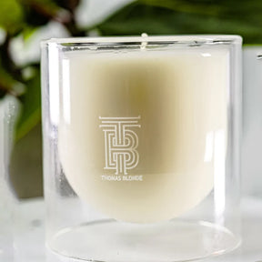 Candle in a clear glass holder with 'THOMAS BLONDE' branding on a blurred natural background