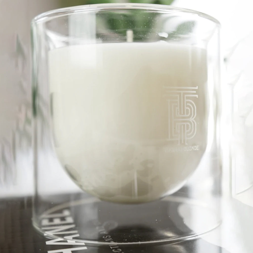 White candle in a glass holder with a brand logo on a reflective surface