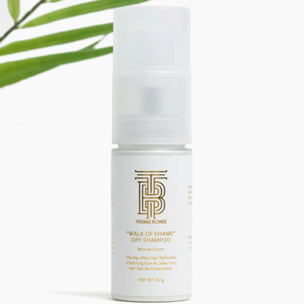 White bottle of 'Walk of Shame' dry shampoo by Thomas Blonde on a white background with green leaves.