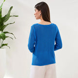 Woman wearing a blue sweater standing in front of a white wall with a plant to the left.