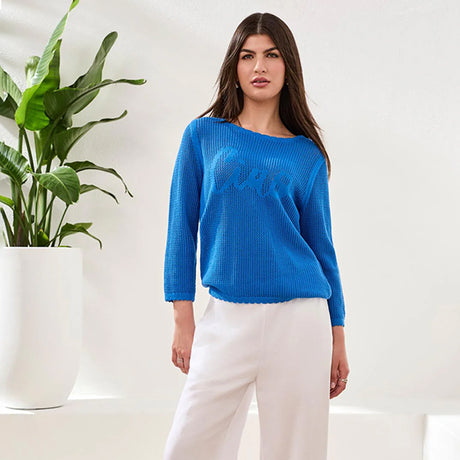 Woman wearing a blue sweater and white pants standing next to a plant indoors.