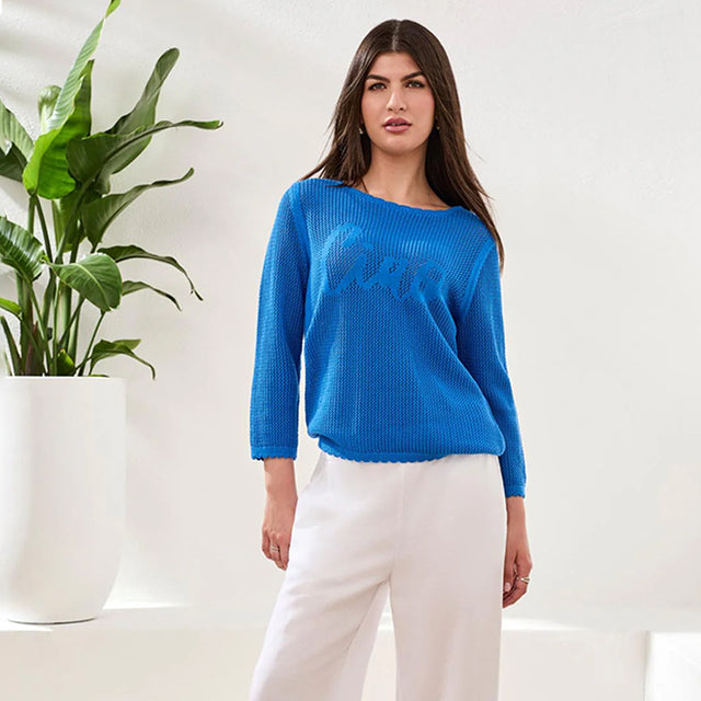Woman wearing a blue sweater and white pants standing next to a plant indoors.