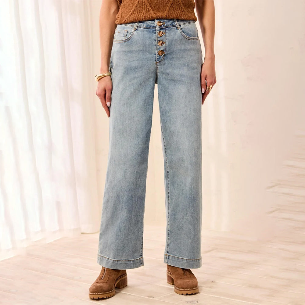 A person wearing light-wash wide-leg jeans with a button-fly waist, paired with a brown knit top and brown suede boots, standing in a softly lit indoor setting with white curtains and light wood flooring.