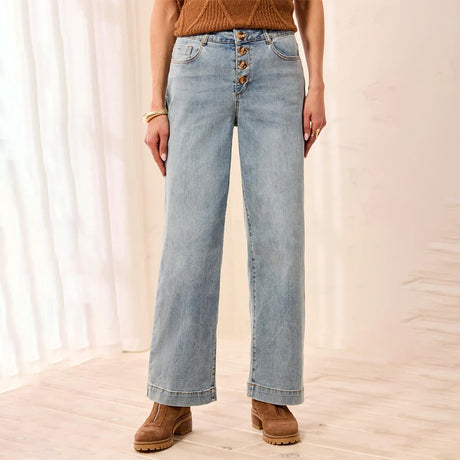 A person wearing light-wash wide-leg jeans with a button-fly waist, paired with a brown knit top and brown suede boots, standing in a softly lit indoor setting with white curtains and light wood flooring.
