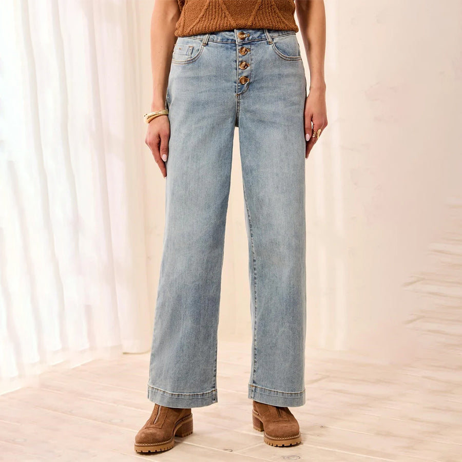 A person wearing light-wash wide-leg jeans with a button-fly waist, paired with a brown knit top and brown suede boots, standing in a softly lit indoor setting with white curtains and light wood flooring.