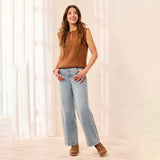 A woman modeling a sleeveless brown cable-knit vest with a zipper neckline, paired with light-wash wide-leg jeans and brown suede boots, standing in a brightly lit room with sheer curtains.
