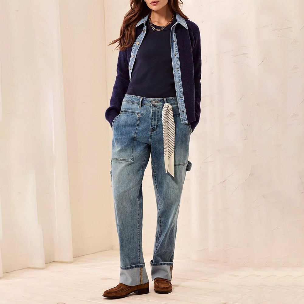 A model wearing light-wash blue straight-leg jeans with a rolled cuff, styled with a navy cardigan, dark top, and brown loafers, standing against a cream-colored textured wall.