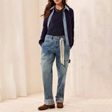 A model wearing light-wash blue straight-leg jeans with a rolled cuff, styled with a navy cardigan, dark top, and brown loafers, standing against a cream-colored textured wall.