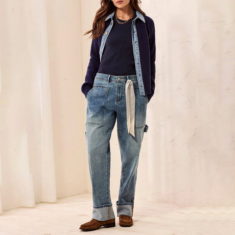 A model wearing light-wash blue straight-leg jeans with a rolled cuff, styled with a navy cardigan, dark top, and brown loafers, standing against a cream-colored textured wall.