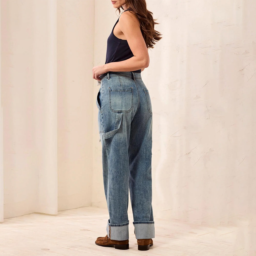 A woman shown from the side and back wearing light-wash blue wide-leg denim jeans with rolled cuffs, a dark sleeveless top, and brown shoes in a softly lit cream-colored room.