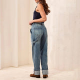A woman shown from the side and back wearing light-wash blue wide-leg denim jeans with rolled cuffs, a dark sleeveless top, and brown shoes in a softly lit cream-colored room.