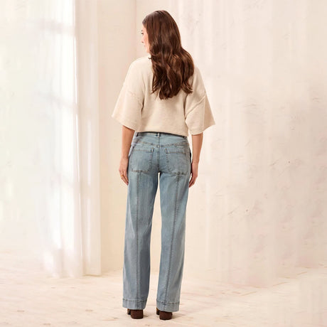 A woman is shown from behind wearing light-wash blue straight-leg jeans and a cream-colored short-sleeve knit top, standing in a softly lit studio with a textured beige wall and sheer white curtains.