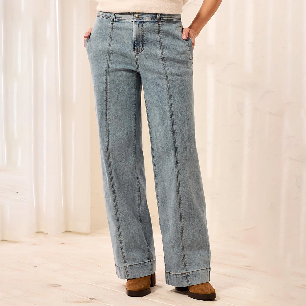A person wearing light blue wide-leg denim jeans with a straight-to-slightly-flared leg, paired with a cream top and brown suede boots, standing in a softly lit indoor setting with light curtains and a pale wood floor.