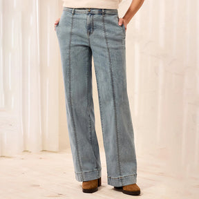 A person wearing light blue wide-leg denim jeans with a straight-to-slightly-flared leg, paired with a cream top and brown suede boots, standing in a softly lit indoor setting with light curtains and a pale wood floor.