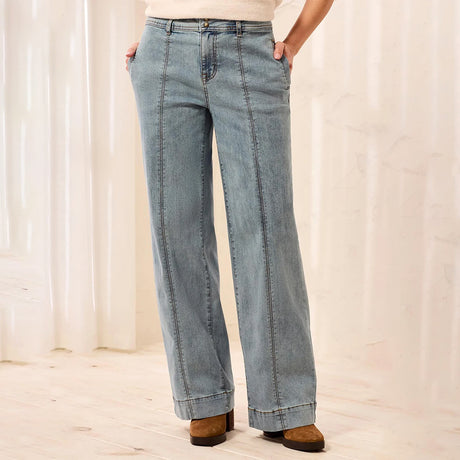 A person wearing light blue wide-leg denim jeans with a straight-to-slightly-flared leg, paired with a cream top and brown suede boots, standing in a softly lit indoor setting with light curtains and a pale wood floor.