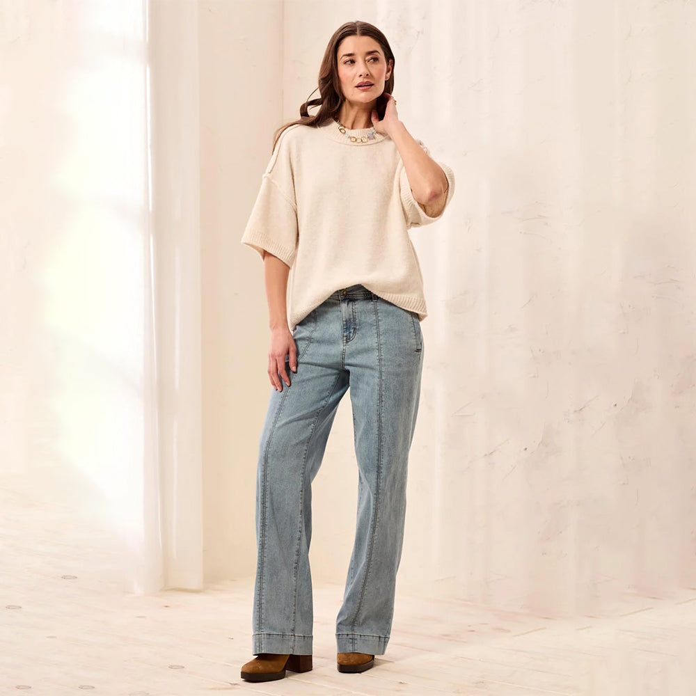 A woman stands in a bright, minimalist studio wearing a cream-colored short-sleeve knit top with a pearl collar detail, light-wash blue wide-leg jeans, and brown leather shoes.