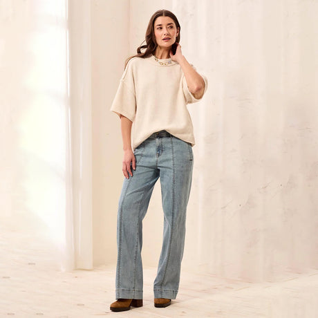 A woman stands in a bright, minimalist studio wearing a cream-colored short-sleeve knit top with a pearl collar detail, light-wash blue wide-leg jeans, and brown leather shoes.