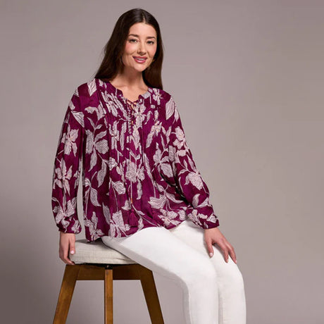 Woman wearing a purple floral blouse sitting on a wooden stool against a tan background