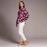 Woman wearing a purple floral blouse and white pants against a gray background