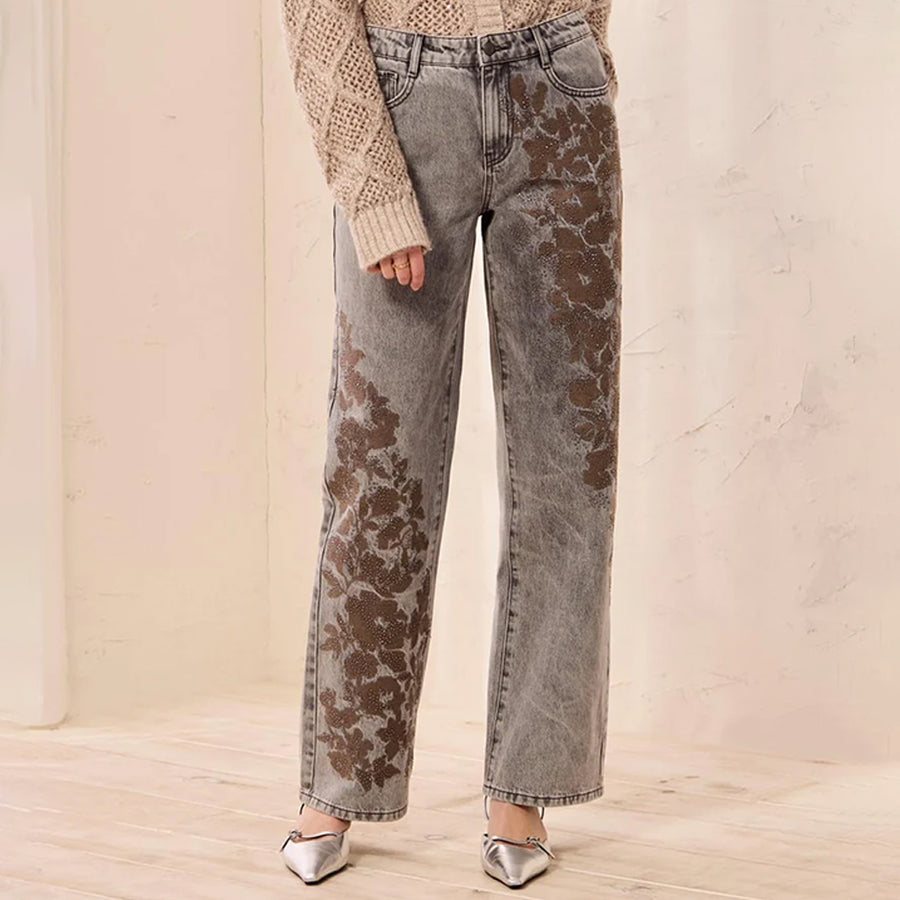 A person wearing gray acid-wash jeans with a brown floral print overlay, styled with a beige knit sweater and silver pointed-toe heels in a softly lit indoor setting.