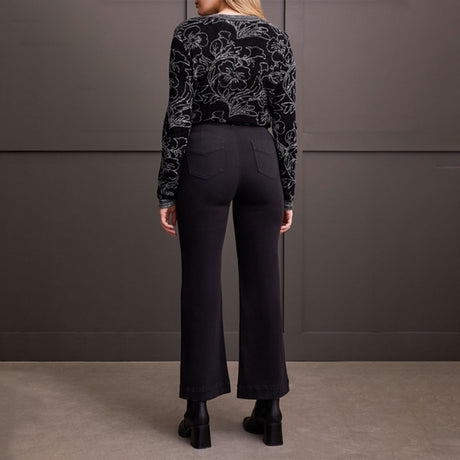 Person wearing a black patterned sweater and black pants against a gray wall.