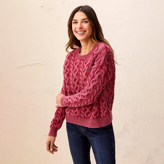 A woman wearing a dusty rose cable-knit sweater and dark blue jeans, standing against a textured off-white wall.